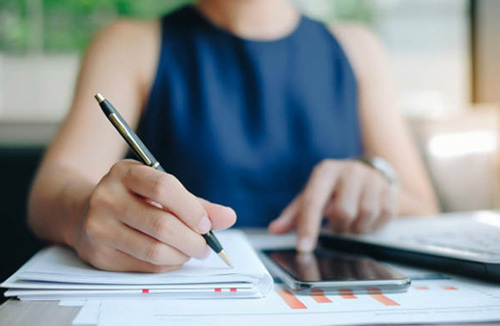 Woman writing in a notebook and using cell phone and laptop
